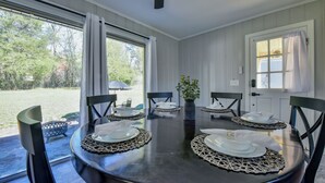 Mesa de comedor preparada para seis personas, con platos blancos y sillas negras, un florero con follaje y vistas a un jardín a través de amplios ventanales.