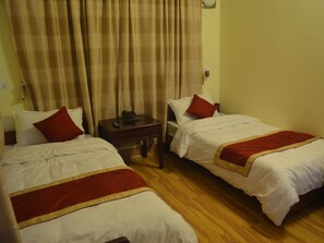 A hotel room with two single beds, each with a red and white bedspread, a wooden nightstand with a telephone, and a curtain with a paisley pattern. - Potala Tourist Home (Kathmandu)