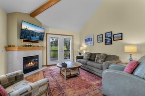 A living room with a fireplace, a television, a sofa, a coffee table, and a view through a glass door to an outdoor area. - New! Eagle Crest Retreat | 3BR Townhome w/ Resort Access—Pet Friendly (Redmond)