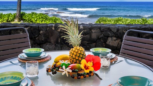 A table set with a tropical fruit arrangement, including a pineapple, banana, and mango, with a view of the ocean and a stone wall in the background. - Easy Walk to Town Ocean Front AC & Wifi - Vrkcv (Kailua-Kona)