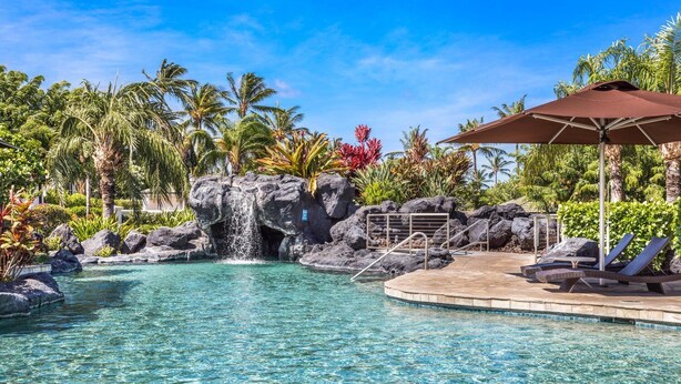 Ein klares Schwimmbecken mit Wasserfall, Liegestühlen und einem schattigen Bereich.