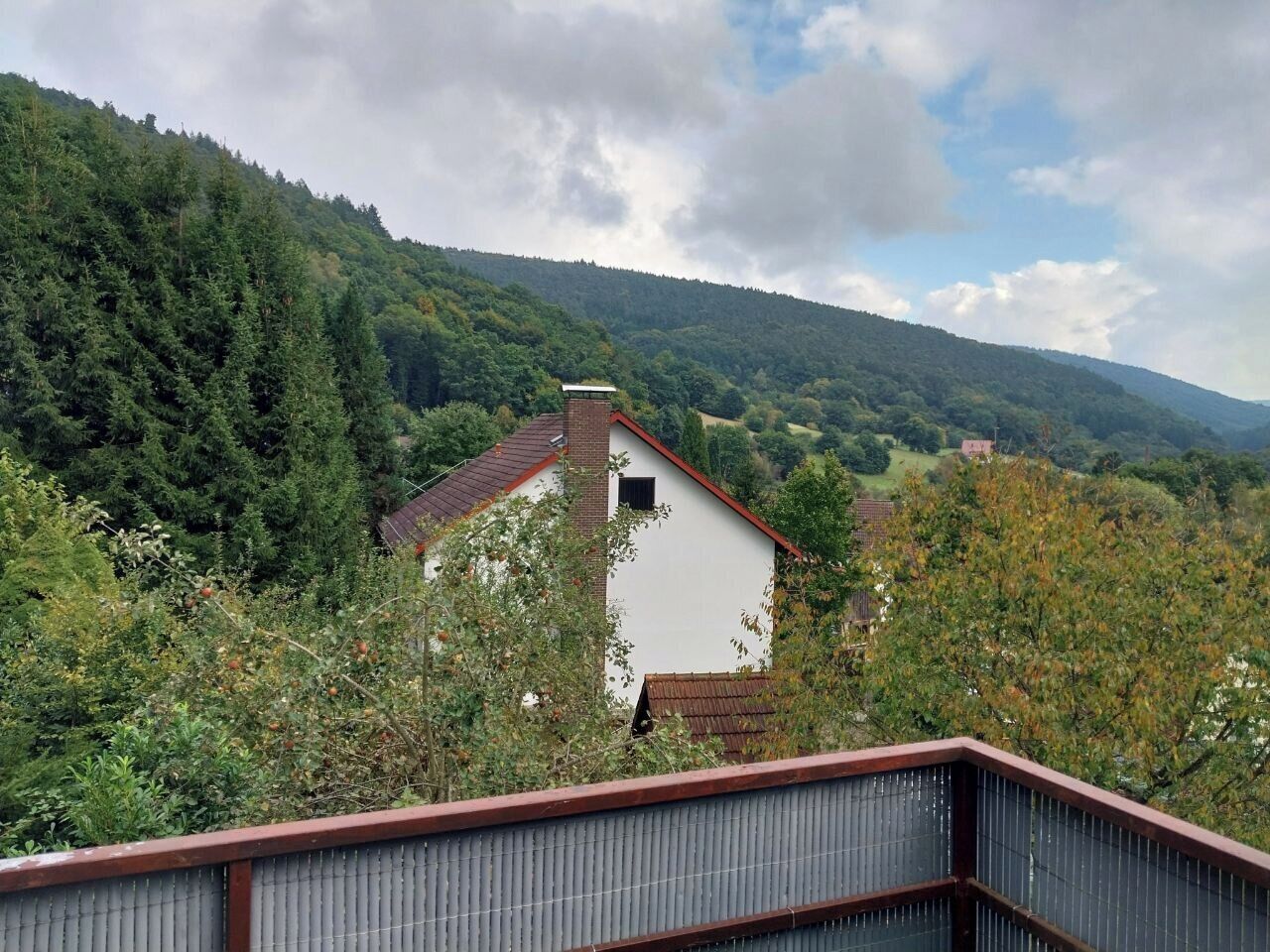 En hvid bygning med rødt tag, omgivet af tæt grøn skov og bakker i baggrunden.