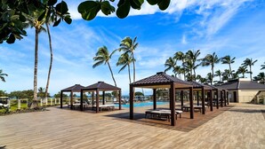Eine Holzterrasse mit Sitzgelegenheiten im Freien und einem Pool, umgeben von Palmen und blauem Himmel.