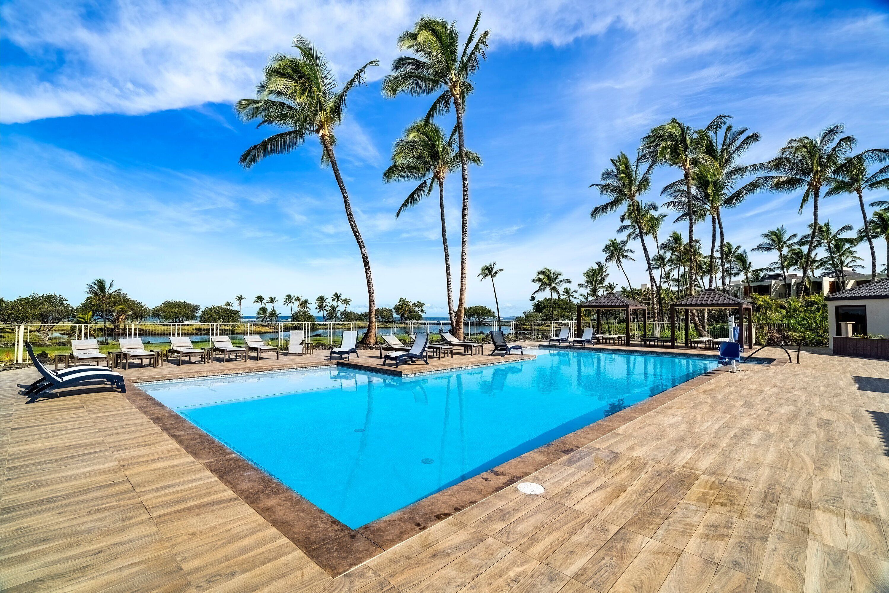 A swimming pool with lounge chairs and palm trees, surrounded by a wooden deck and a view of the ocean.