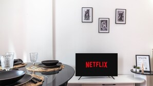 A modern dining area with a glass table, black plates, and clear glasses. A flat-screen TV displaying "NETFLIX" is mounted on a white cabinet. The wall features framed pictures and a welcome sign.