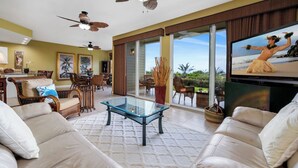A spacious living room with a glass coffee table, comfortable seating, and a large TV screen displaying a beach scene. - Updated Townhome Ocean View Private BBQ & Lanai - Vrkcv (Kailua-Kona)