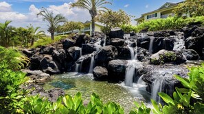 Une cascade naturelle formée par des rochers, avec une eau claire, entourée d’une végétation luxuriante et un bâtiment visible au loin.
