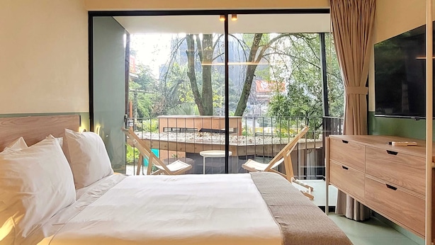 A hotel room with a large bed, a wooden dresser, a television, and a balcony with a view of trees and a building. - w Attractive 2BR w Balcony in Poblado (Medellín)
