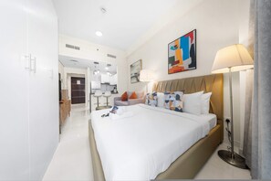 A modern hotel room with a large bed, a bedside lamp, a painting, and a view into the kitchen area. - Stayshort - Studio in JVC Sleeps 2 With City Views (Dubai)