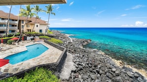 A pool area with a view of the ocean, surrounded by a stone wall and palm trees. - Ocean Front Fantastic Views Corner Unit - Vrkcv (Kailua-Kona)