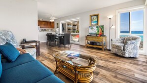 A living room with a sofa, a wicker coffee table, a chair, and a view of the ocean through a large window. - Ocean Front Fantastic Views Corner Unit - Vrkcv (Kailua-Kona)