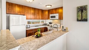 A kitchen with wooden cabinets, a white refrigerator, a microwave, a sink, and a countertop with fruits. - Ocean Front Fantastic Views Corner Unit - Vrkcv (Kailua-Kona)