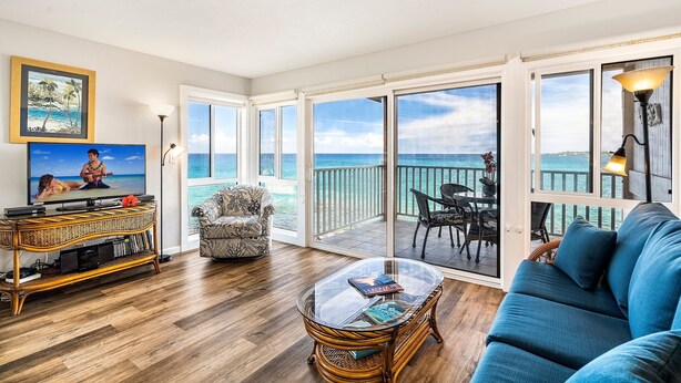A spacious living room with a large TV, a blue sofa, a wicker coffee table, and a view of the ocean through large sliding doors. - Ocean Front Fantastic Views Corner Unit - Vrkcv (Kailua-Kona)