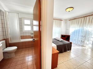 Une salle de bain comprenant des toilettes, un lavabo et un espace douche. La pièce est dotée d’un carrelage au sol et d’une fenêtre avec des rideaux.