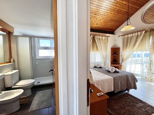 Une chambre d’hôtel avec un plafond en bois, un lit recouvert d’une couette blanche, une table de chevet avec une lampe, et une vue sur la mer par de grandes fenêtres.