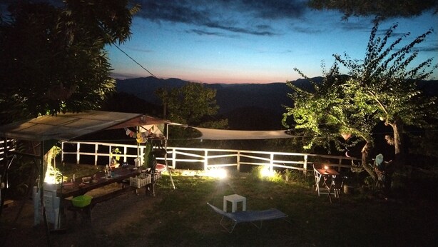 A nighttime outdoor area with a covered seating area, a table with chairs, and a view of distant mountains. - In the heart of the forest stands Pachamama, 900 m above sea level detached cottage (Bajardo)