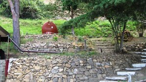 A stone wall with a metal staircase leading up to a red spherical object. - In the heart of the forest stands Pachamama, 900 m above sea level detached cottage (Bajardo)