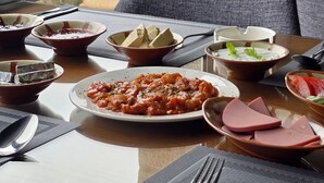 A dining table with various dishes, including pasta, meat, and a bowl of soup, set on a mat with a view of the outdoors through a window.