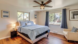 Une chambre à coucher comprenant un lit, une table de chevet, un ventilateur de plafond, deux fenêtres avec des rideaux et une chaise.