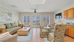 A spacious living area with a sofa, chairs, a coffee table, and a dining table. There is a ceiling fan, a television, and a view of the outdoors through the glass doors. - Blue Dolphin, Oceanfront Views! Sandbridge Home (Virginia Beach)