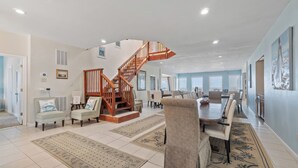 A spacious living area with a wooden staircase, a dining table with chairs, and a large window providing natural light. - Blue Dolphin, Oceanfront Views! Sandbridge Home (Virginia Beach)