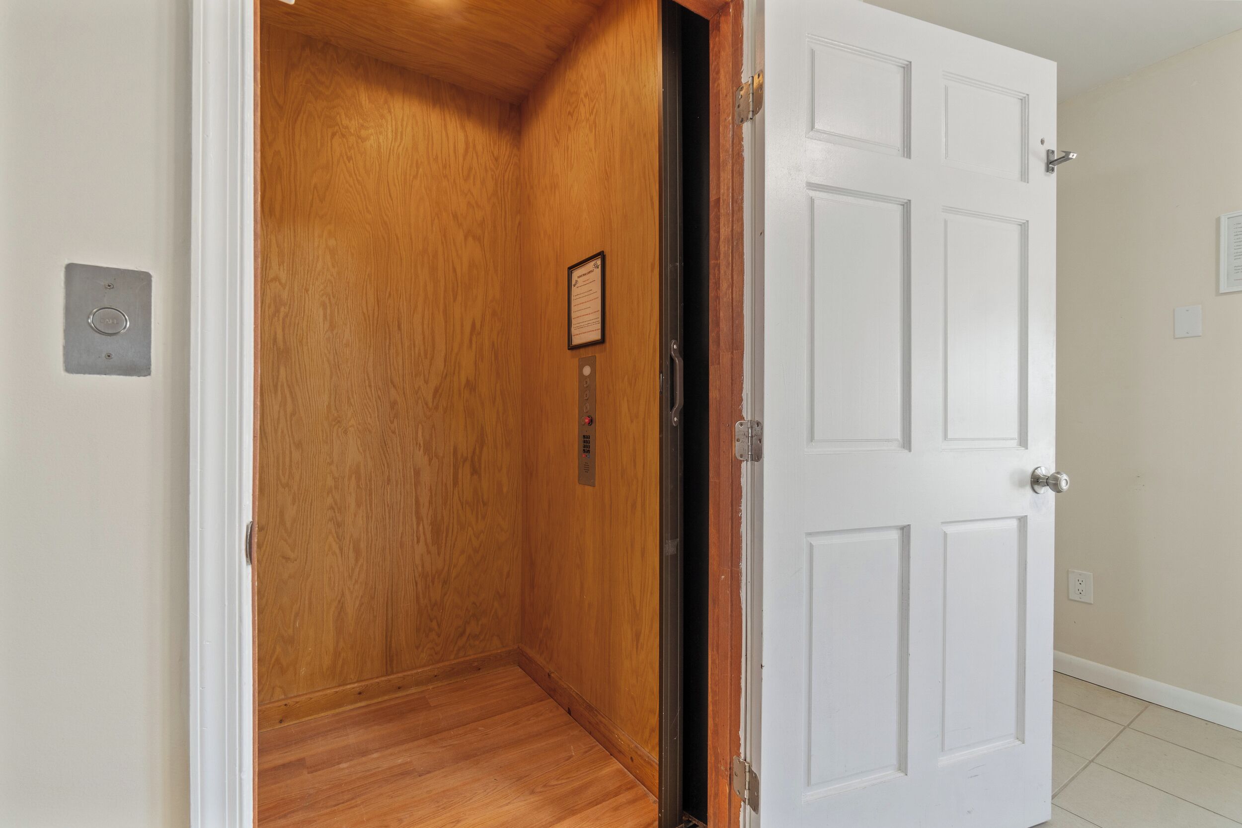 Un couloir avec une porte en bois et une porte blanche, toutes deux dotées de poignées apparentes et d’une petite plaque sur la porte en bois.