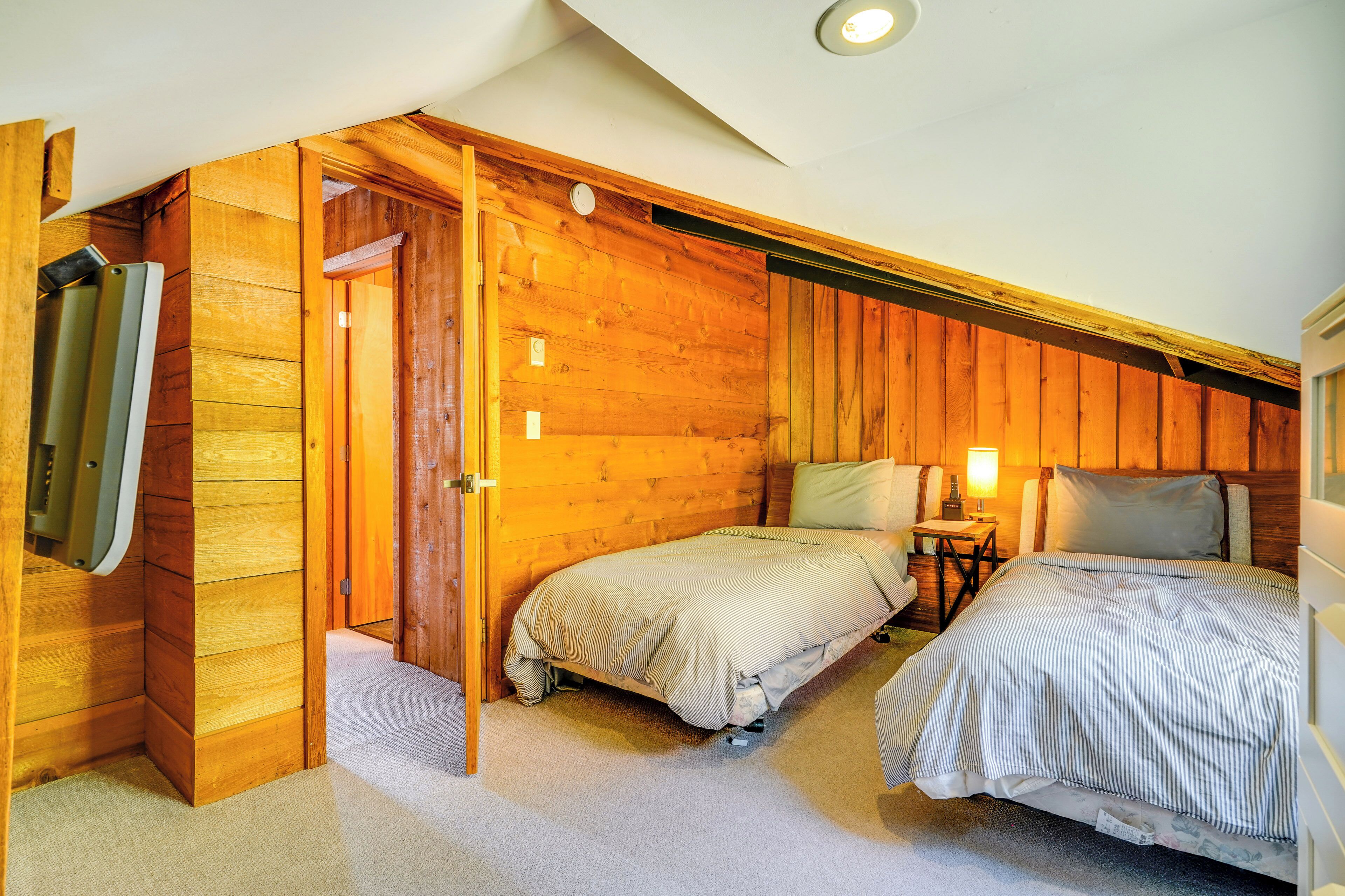 Un dormitorio con dos camas, paredes de madera, una mesita de noche con lámpara y una puerta que da a otra habitación.