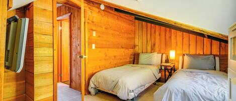 Un dormitorio con dos camas, paredes de madera, una mesita de noche con lámpara y una puerta que da a otra habitación.