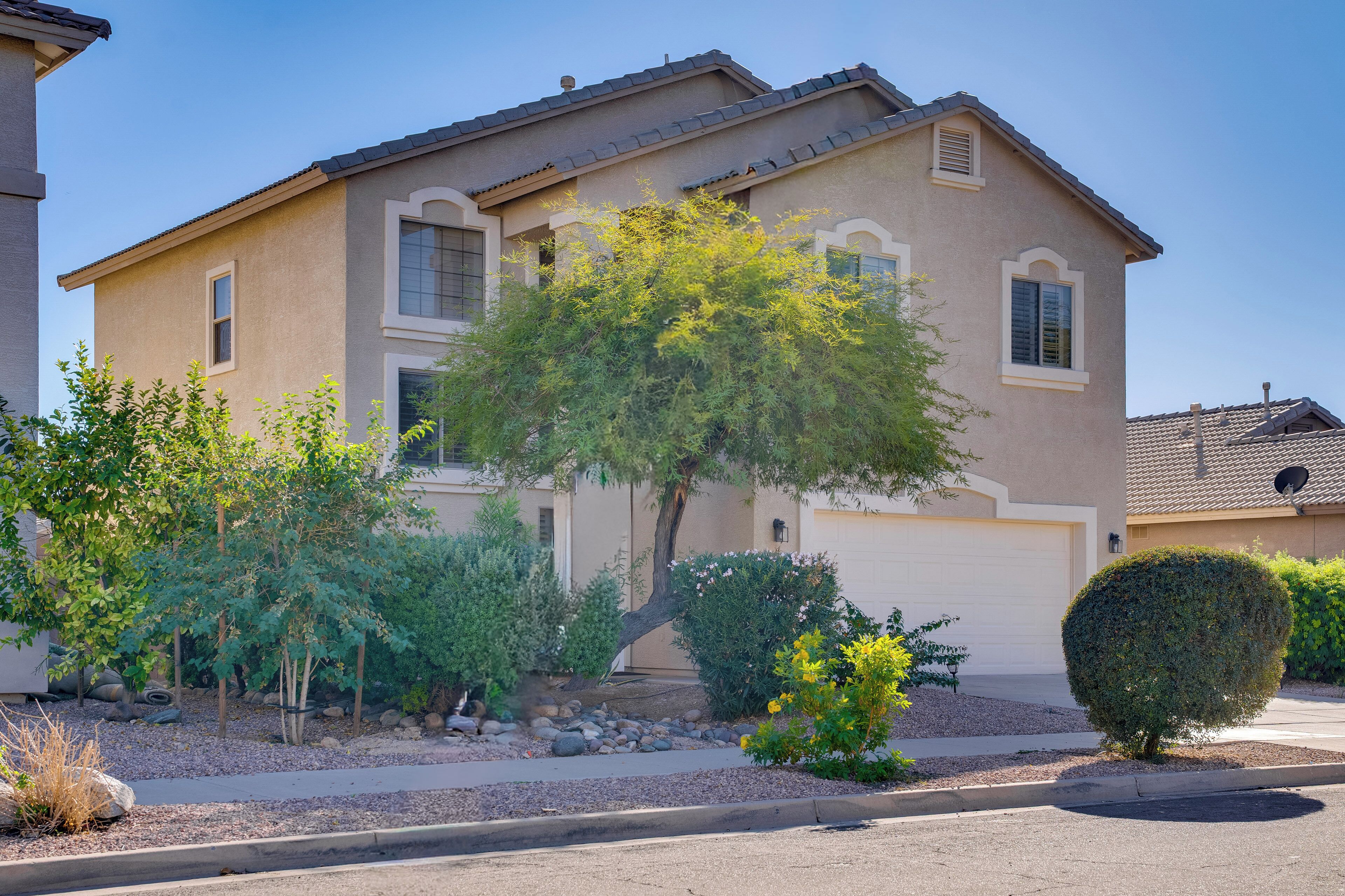A two-story house with a beige exterior, a garage, and a well-maintained front yard with various shrubs and a tree.