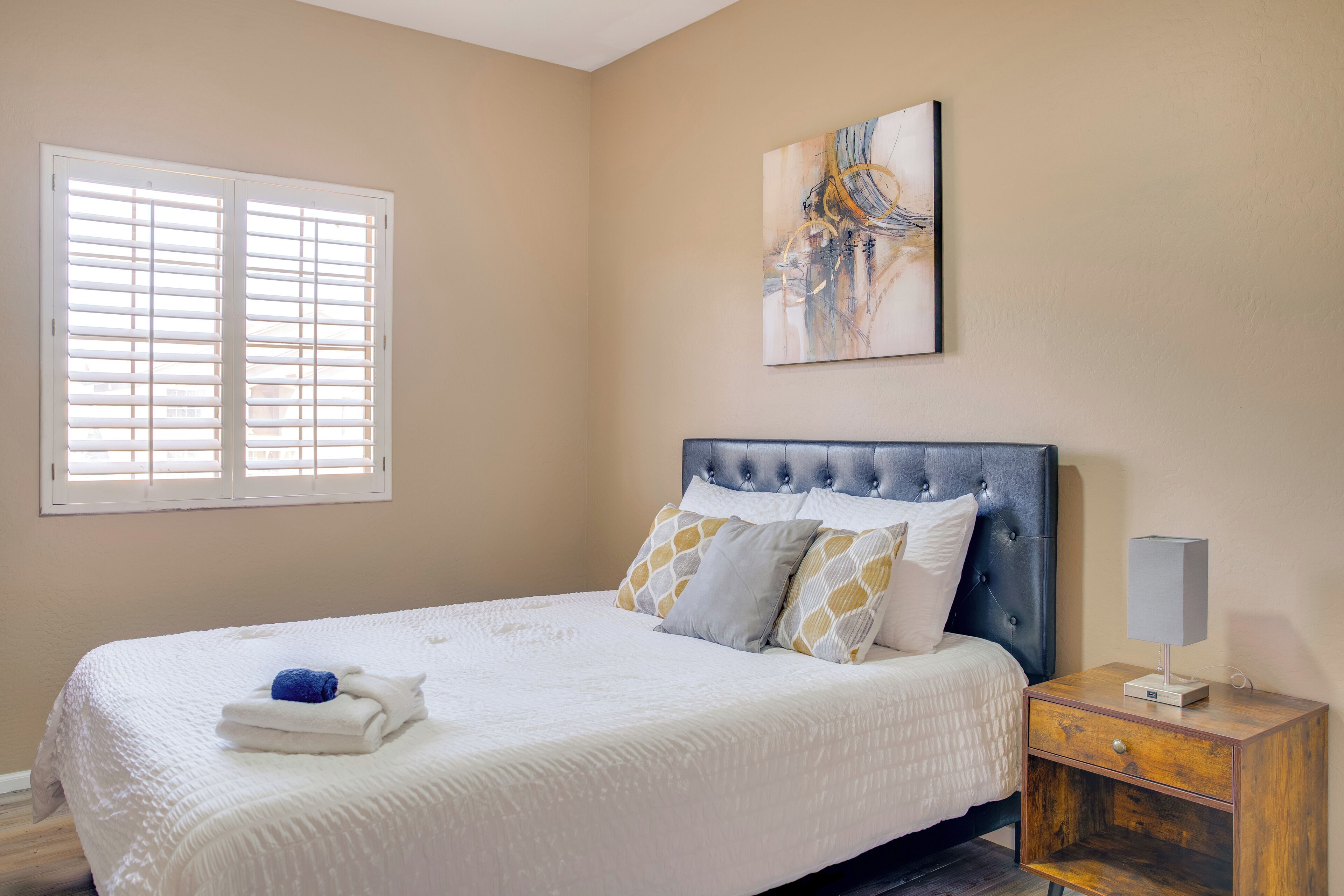 Quarto com cama, mesa de cabeceira, luminária, janela com persianas e um quadro na parede.