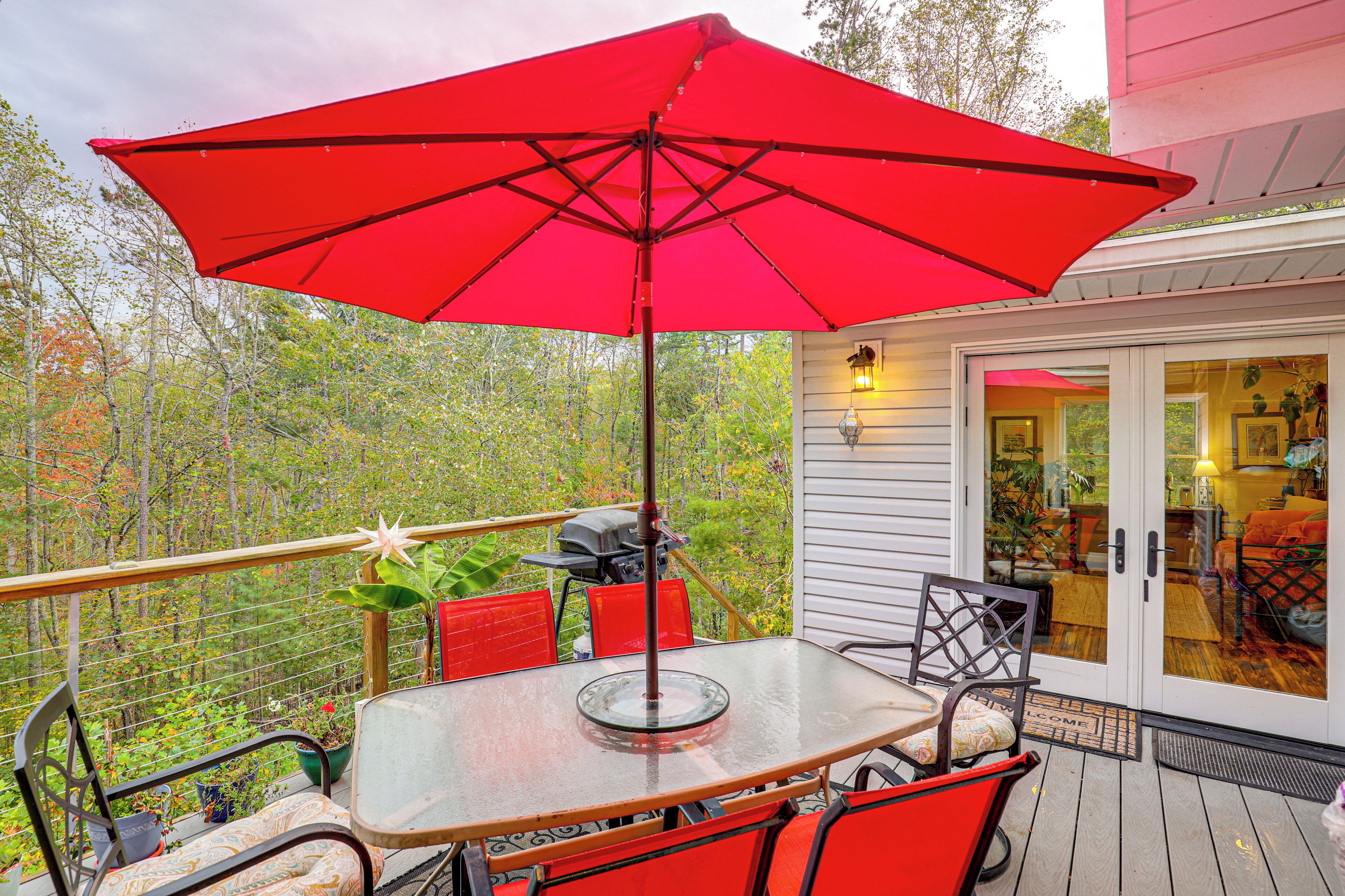A glass-top table with red chairs and a red umbrella on a wooden deck.