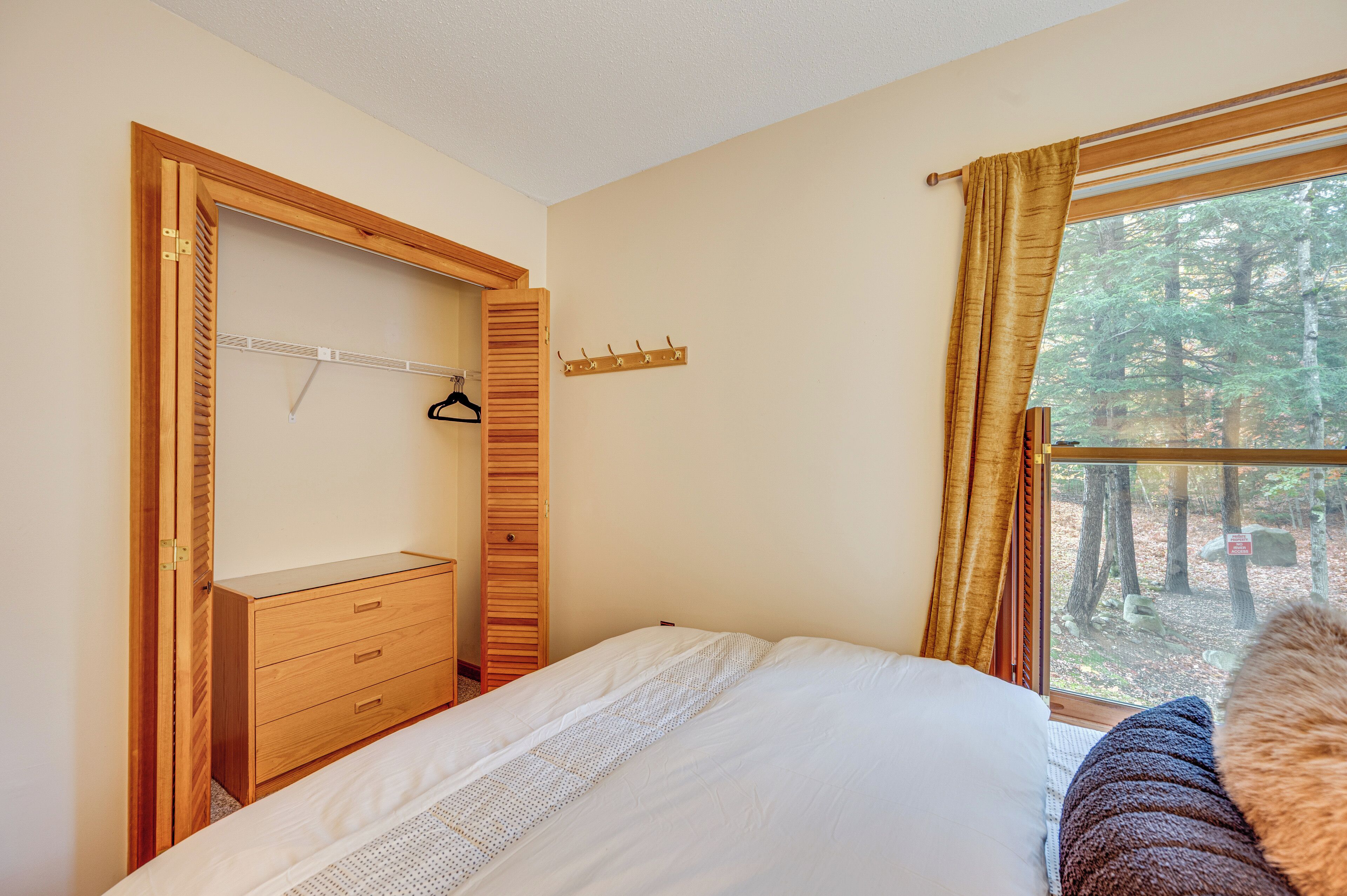 Ein Schlafzimmer mit Holzschrank, Bett, Fenster mit Blick auf einen Wald und einer Garderobe.