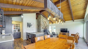 A spacious kitchen with a large island, wooden dining table, and chairs, a stone fireplace, and a television in a modern living area. - 2 Mi to Beech Mtn Resort: Family Home w/ Hot Tub! (Banner Elk)