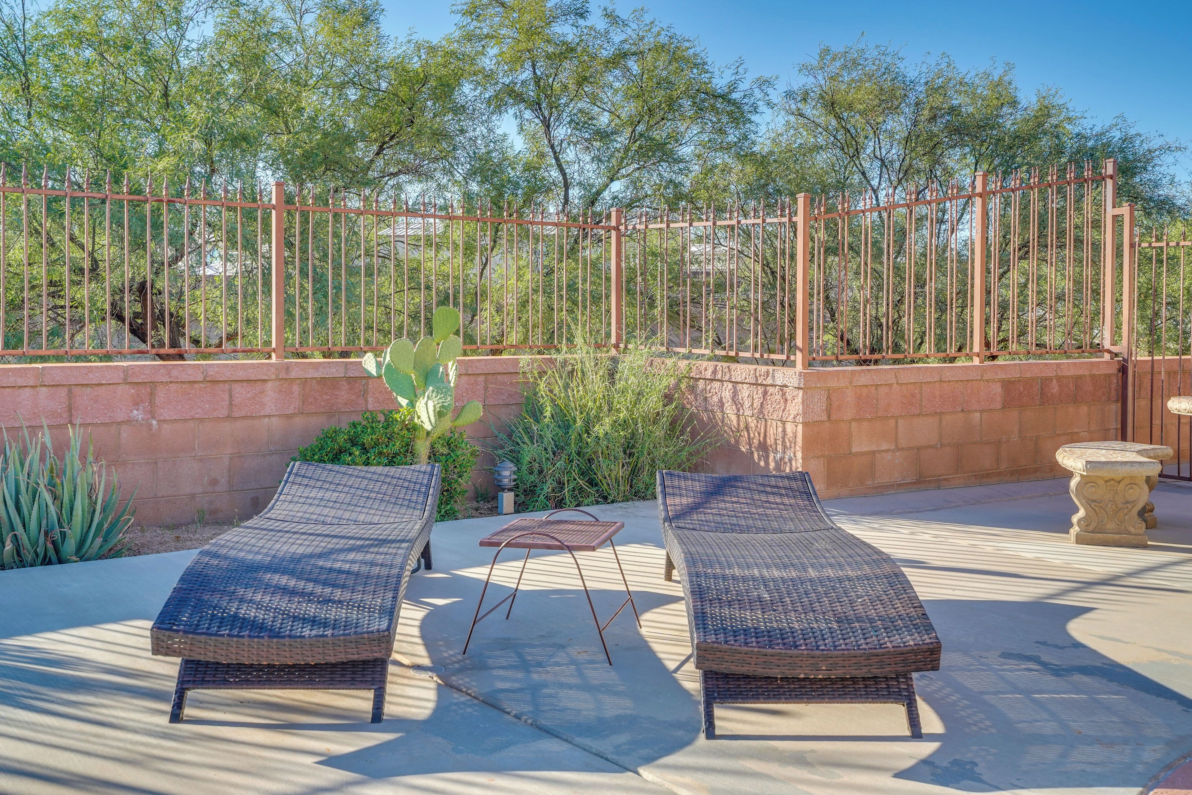 An outdoor seating area with two wicker lounge chairs and a metal table, surrounded by a brick wall and a metal fence, with trees in the background.