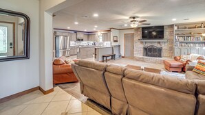 A spacious living area with a leather sofa, a fireplace, a television, and a kitchen in the background. - Tulsa Area Home w/ Sunroom: Near Parks & River! (Bixby)