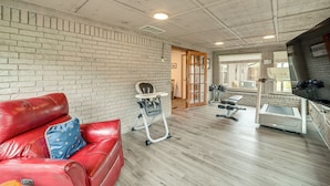 A room with a brick wall, a red leather sofa, a baby rocker, a treadmill, and a television. - Tulsa Area Home w/ Sunroom: Near Parks & River! (Bixby)