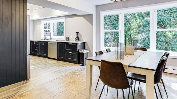 A modern kitchen with dark cabinetry, a wooden floor, and a dining area with a table and chairs.