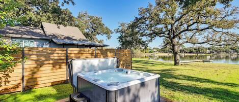 Un jacuzzi en un patio trasero con cerco de madera, una casa de ladrillo y vista al lago.