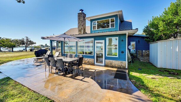 A two-story house with a patio area featuring a table and chairs, a stone chimney, and a view of a body of water.