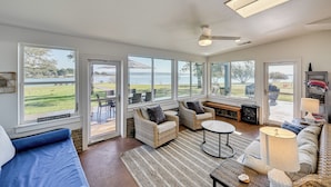 A spacious living room with large windows, wicker furniture, a ceiling fan, and a view of the outdoors.
