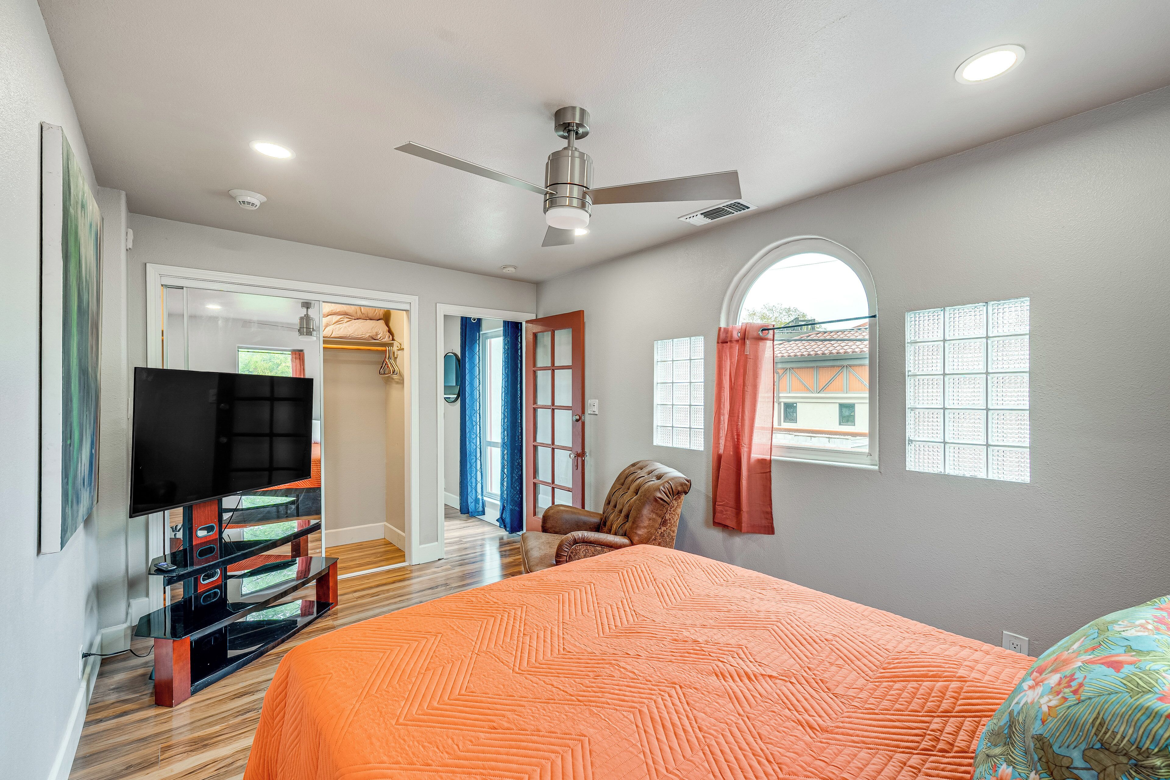 Un dormitorio con una cama, un televisor sobre un mueble bajo, un ventilador de techo y una ventana con cortinas.