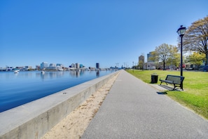 A waterfront walkway with a bench and trash can, leading to a calm body of water with boats and buildings in the background. - Steps to Elizabeth River & Dtwn! Portsmouth Condo (Portsmouth)
