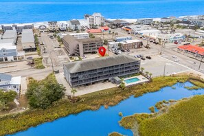 Une vue aérienne d’une ville côtière, où un bâtiment remarquable est signalé par une épingle rouge, entouré de bâtiments résidentiels et commerciaux, d’une plage et d’une étendue d’eau.