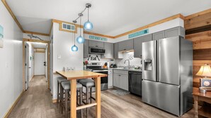 A modern kitchen with stainless steel appliances, a wooden dining table with bar stools, and pendant lighting. - Walkin on Sunshine! Marshside Inn 309 (Murrells Inlet)