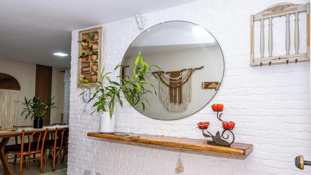 Een eethoek met een houten tafel en stoelen, een witte bakstenen muur, een ronde spiegel en een plank met decoratieve objecten.