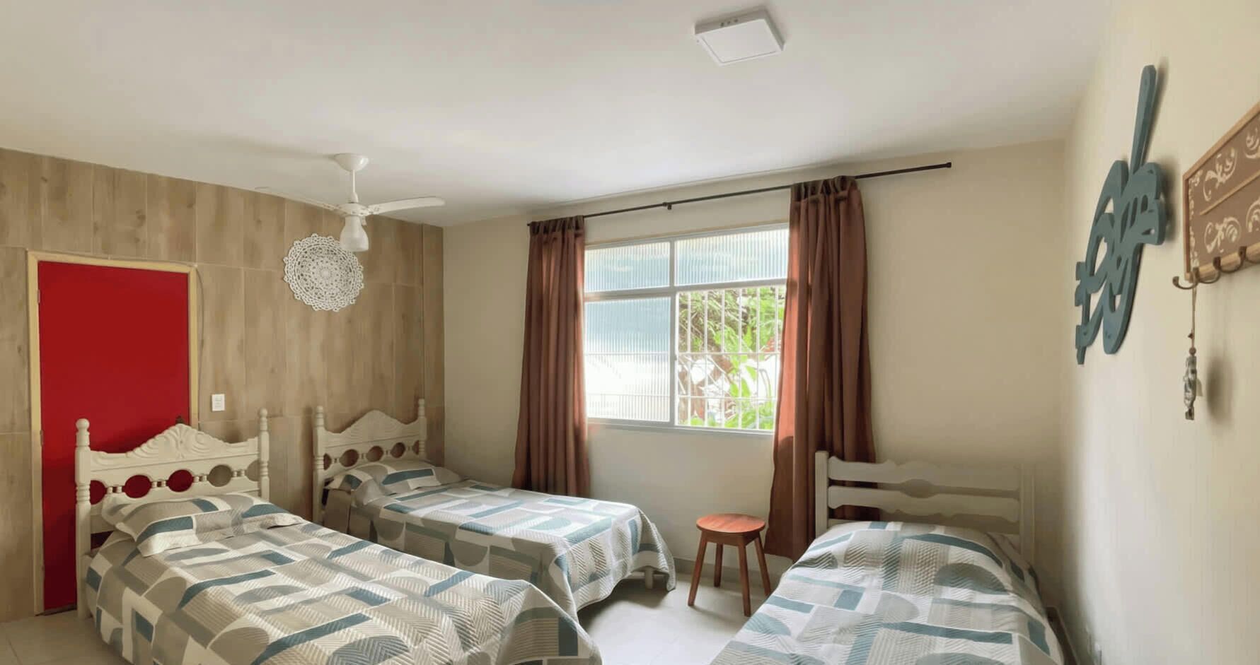 A room with three single beds, a ceiling fan, a window with curtains, and a small stool.