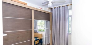 Ein Zimmer mit Schiebetürenschrank, Deckenventilator, Fenster mit Vorhängen und einem kleinen Tisch mit Stuhl.