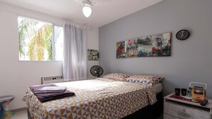 Une chambre à coucher avec un lit, une horloge murale, un tableau, un ventilateur et une fenêtre avec des rideaux.