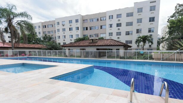 Une piscine dont les bords sont recouverts de carreaux, entourée d’une clôture blanche et située à côté d’un immeuble à plusieurs étages doté de balcons.