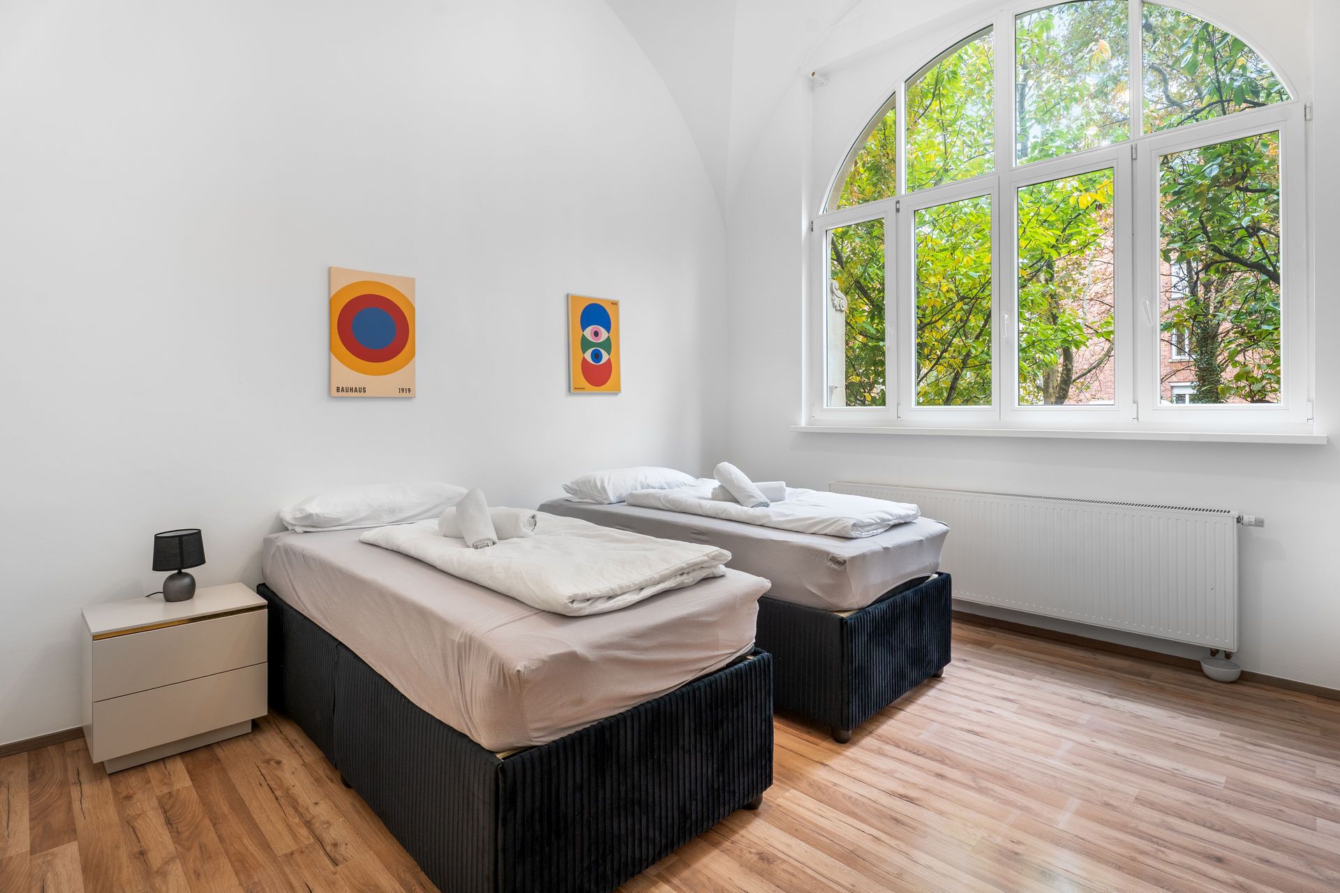 Ein Schlafzimmer mit zwei Betten, einem Nachttisch, einer Lampe und einem großen Fenster mit Blick auf Bäume.